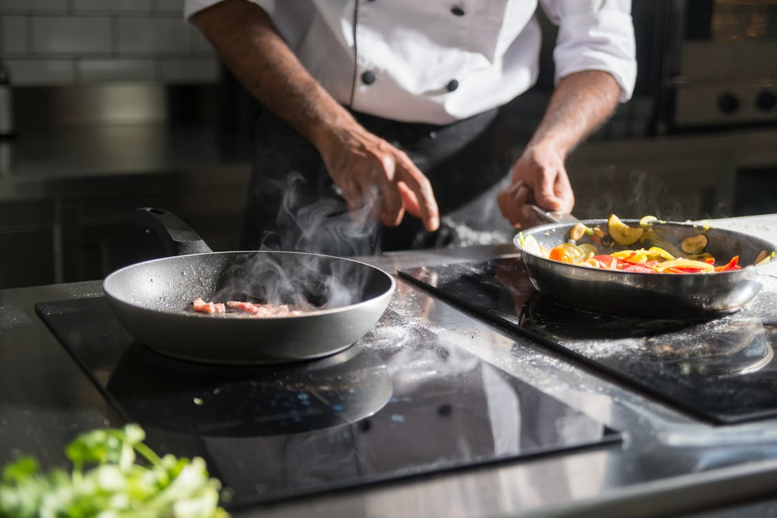 Person cooking at a stovetop with two pans of sizzling vegetables and fresh herbs in a kitchen