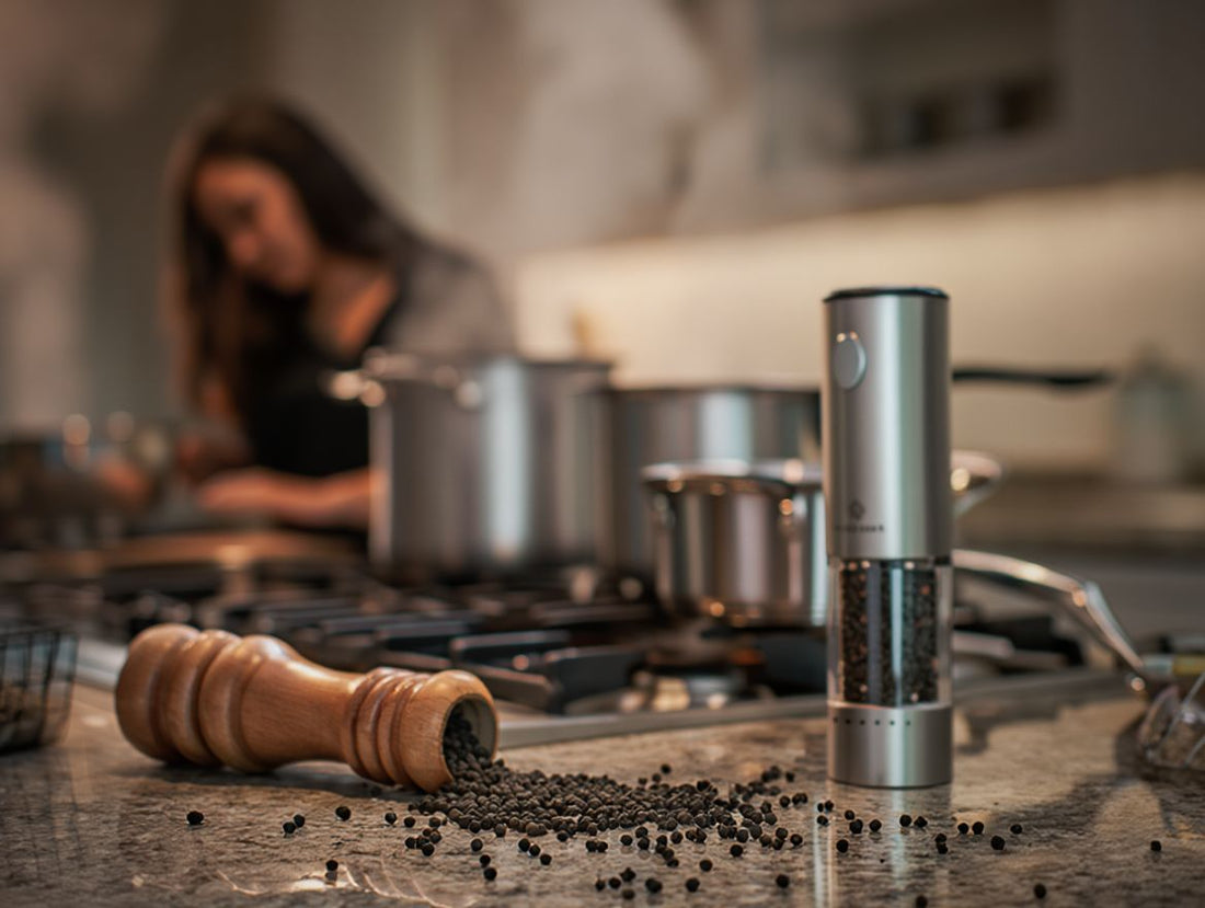 Electric pepper grinder standing next to a tipped-over wooden manual grinder with scattered peppercorns on a kitchen counter