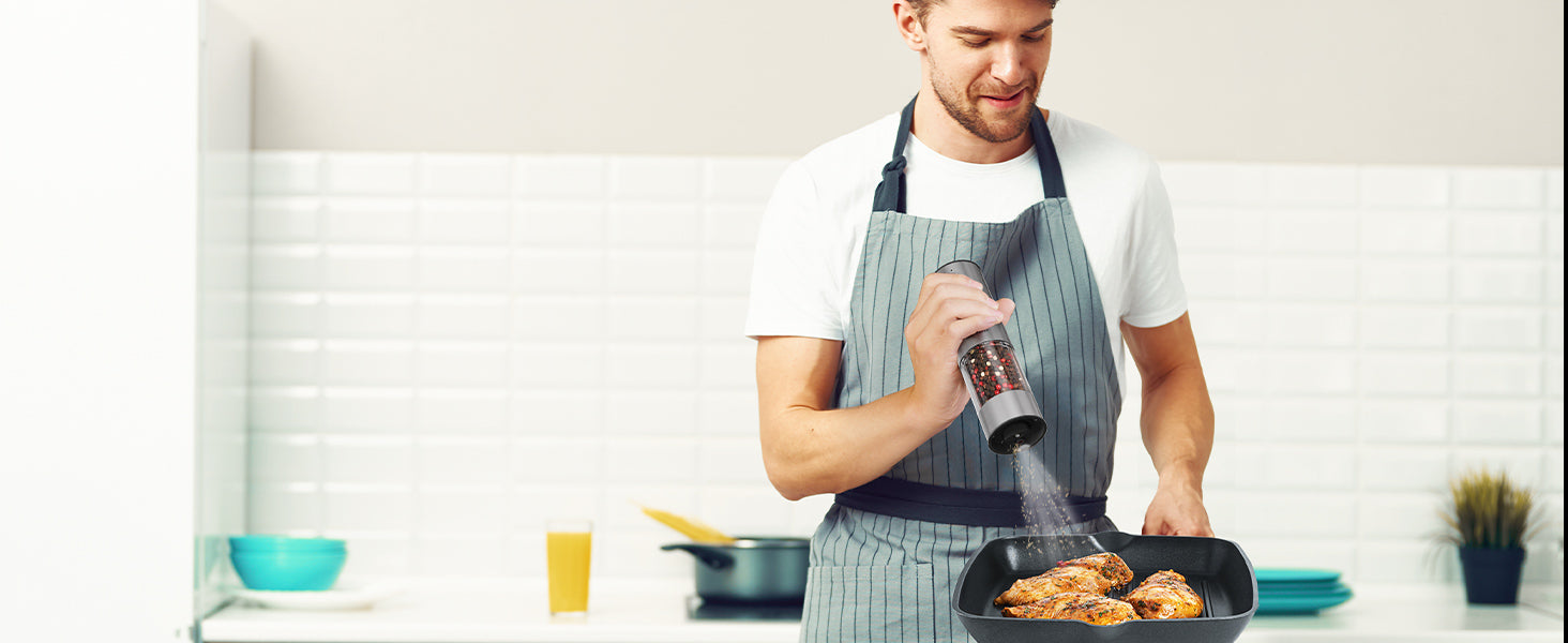 Man in a kitchen wearing an apron, holding a salt and pepper grinder and a pan with food.