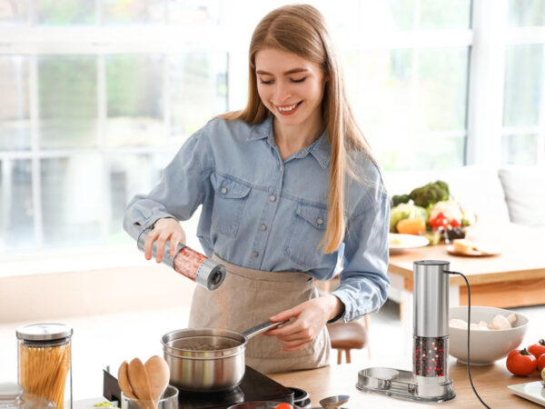 Woman using Vivosparks electric salt and pepper shaker over a pot in a bright kitchen, with charging base and stainless steel shaker set visible on the counter